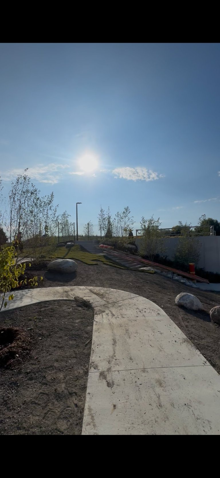 Outdoor construction site with concrete pathways, dirt terrain, landscaping plants, and bright blue sky with sun shining overhead