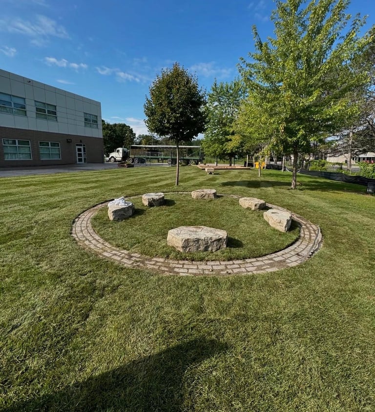 Outdoor school courtyard with a circular seating area made of large boulders and brick pathway on green lawn